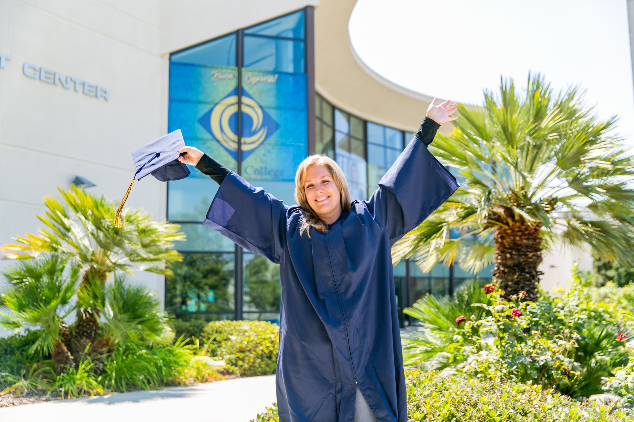 A woman, Emily Costello, in graduation garb raises her mortarboard above her head on the Cypress College campus.