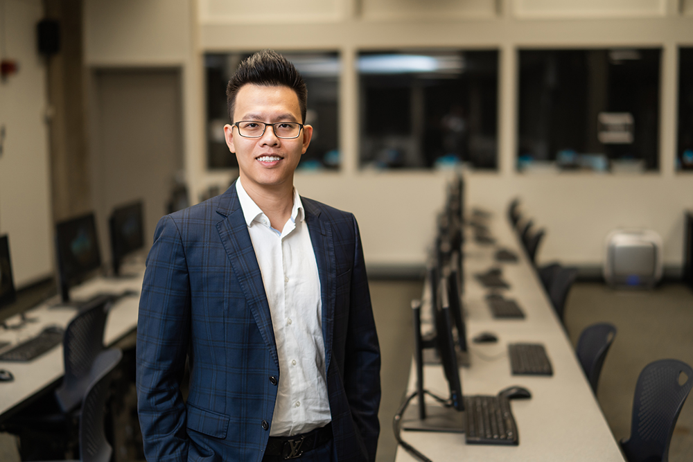 A man in a suit stands before a row of computers as he smiles at the camera.