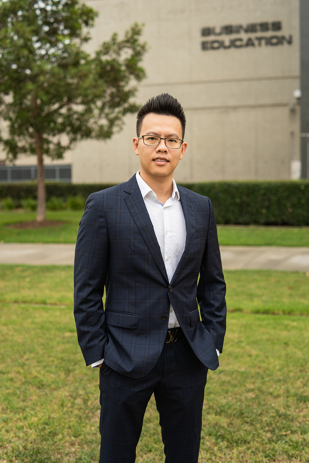 A man with dark hair and wearing a suit with no tie smiles in front of the Cypress College Business Education building on campus.