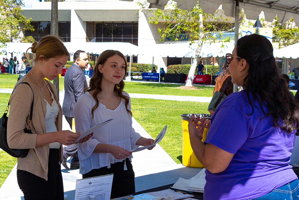 Two students in professional attire speaks to a company representative at an information table on campus.