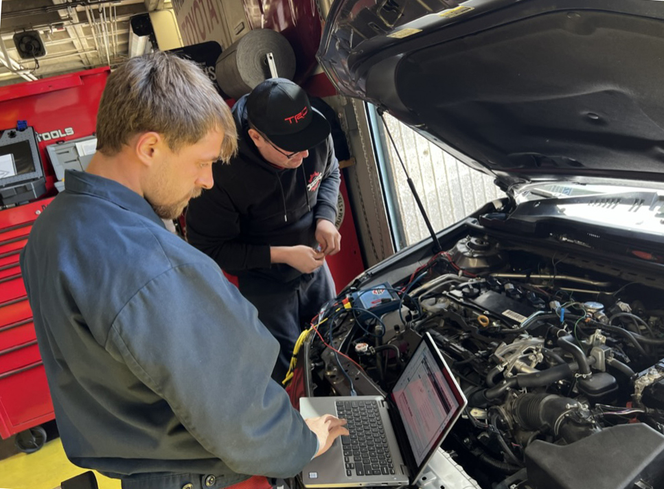 Two men look under the hood of a car while checking diagnostics on a laptop.