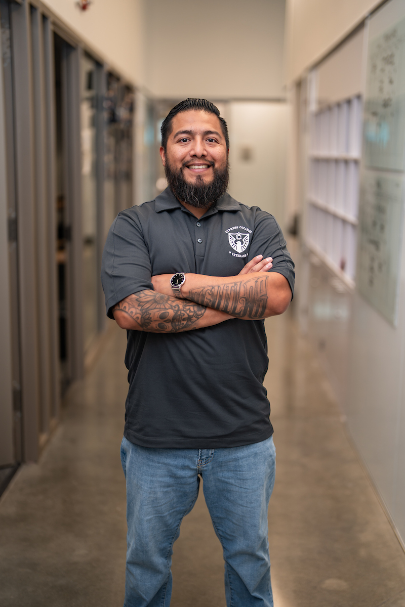 A man with black hair and bear folds his arms and smiles as he stands in a hallway
